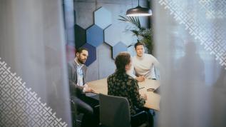 Neuvottelupöydän ääressä istuu henkilöitä. Neuvotteluhuoneen sinertävällä päätyseinällä on kuusikulmioisia kohokuvioita. Osan neuvottelijoista peittää etualalla olevat läpikuultavat verhot.