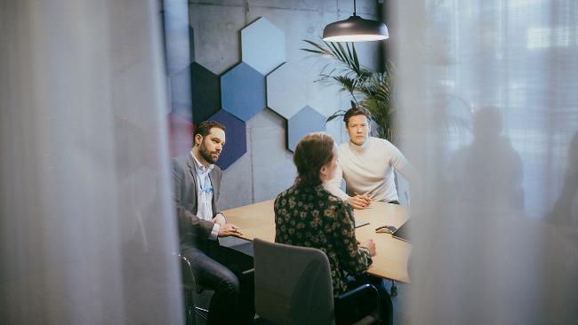 Neuvottelupöydän ääressä istuu henkilöitä. Neuvotteluhuoneen sinertävällä päätyseinällä on kuusikulmioisia kohokuvioita. Osan neuvottelijoista peittää etualalla olevat läpikuultavat verhot.