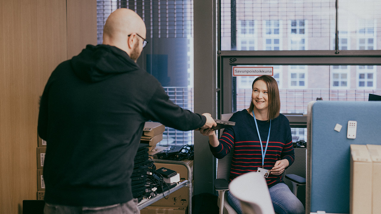 IT-laite menee kierrätykseen. Mustahupparinen mies  on selin, ja hän ojentaa läppärinsä naiselle, jolla on päällään sininen puserossa, jossa on punaisia raitoja. Nainen ojentaa kätensä ottaakseen laitteen itselleen. Metallisessa kärryssä on pino käytöstä poistettuja vanhoja läppäreitä.