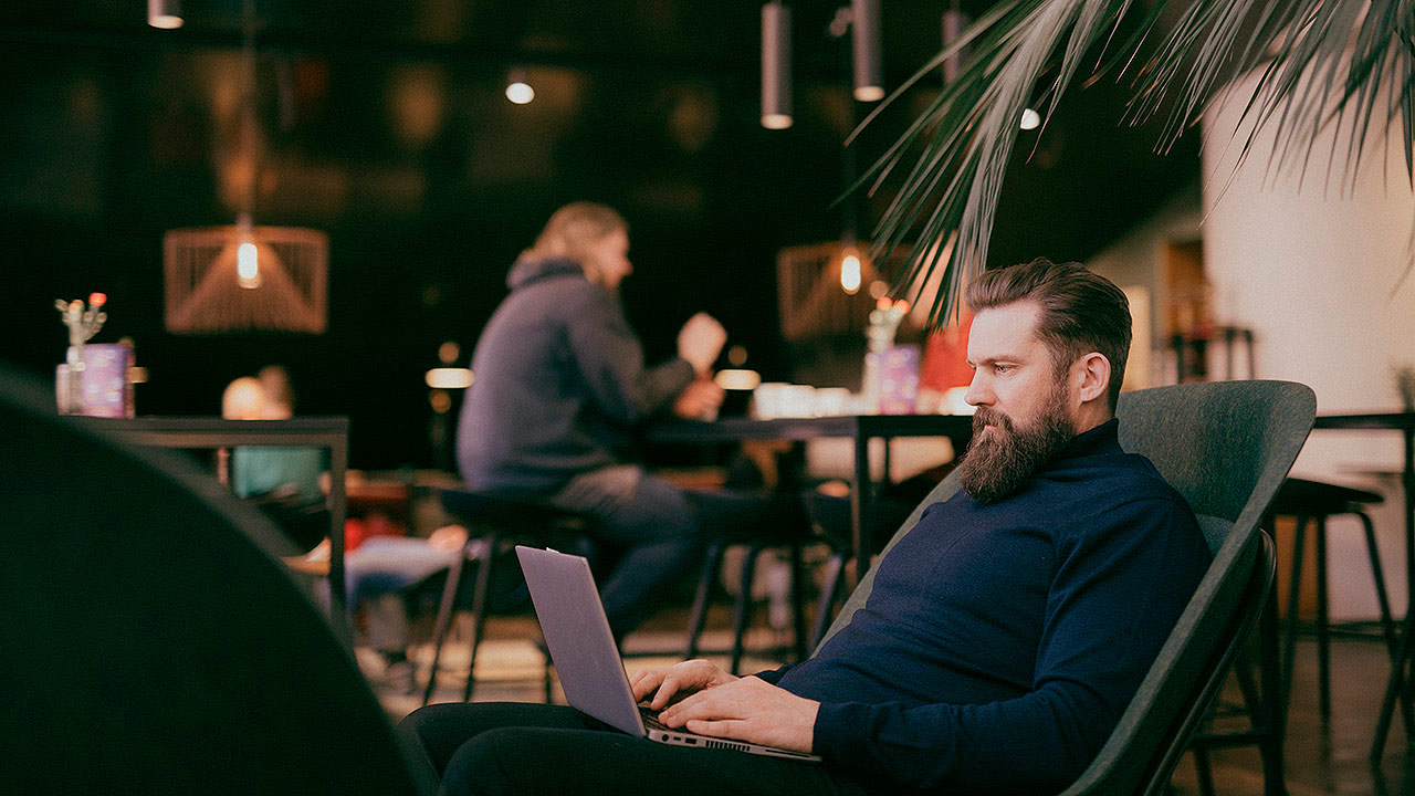 Etualalla istuvalla ruskeatukkaisella, sinipuseroisella ja parrakkaalla miehellä on sylissään läppäri, jota hän katselee. Mies istuu tuolissa sivuttain ja taustalla näkyy muita ihmisiä. Tila muistuttaa tunnelmallista kahvilaa.