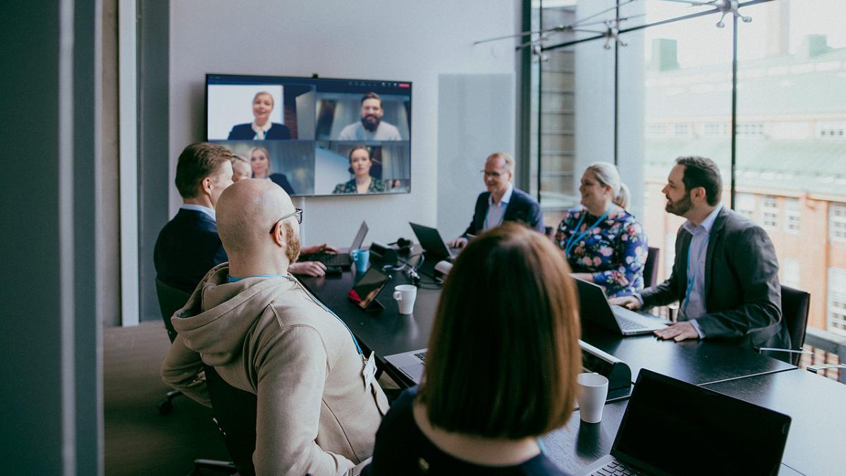 Ihmisiä kokoustamassa pöydän ympärillä, osa on etänä ja heidän kuvansa näkyy skriinillä, TV:n kuvaruudulla.